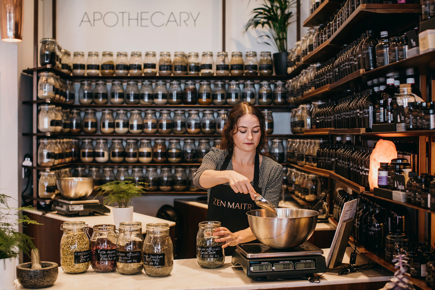 Woman working in Zen Maitri with shelves stocked with jars labelled 'Apothecary'.