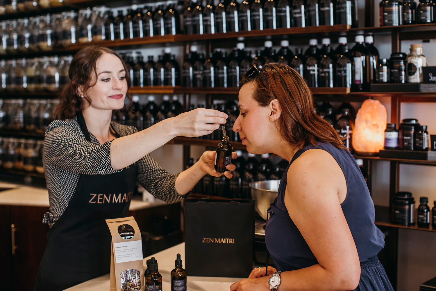 Woman receiving a product sample from another woman in a store setting with shelves of products.